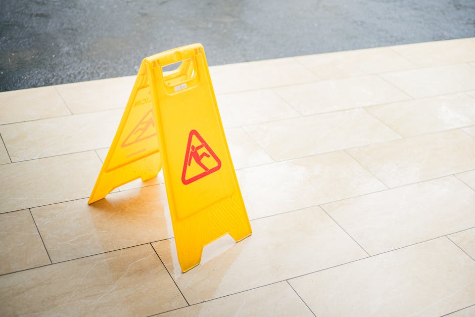 A bright yellow, A-frame wet floor caution sign with red warning symbols, placed on a smooth beige tiled outdoor surface. The sign indicates a slippery area, likely due to recent watering or cleaning,