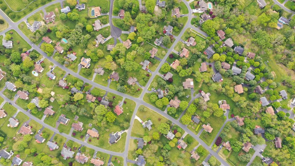 An aerial view of a residential neighborhood showing numerous houses surrounded by lush green trees, with winding roads and pathways weaving through the estate. Some gardens feature well-maintained la