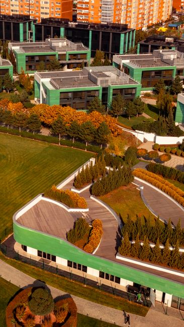 An aerial view of a residential neighborhood showing numerous houses surrounded by lush green trees, with winding roads and pathways weaving through the estate. Some gardens feature well-maintained la