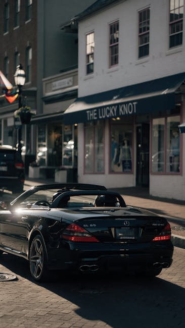 A sleek black convertible sports car is parked on a cobblestone street in front of a building with large windows and a sign reading 'The Lucky Knot.' The car's polished surface reflects nearby sunligh